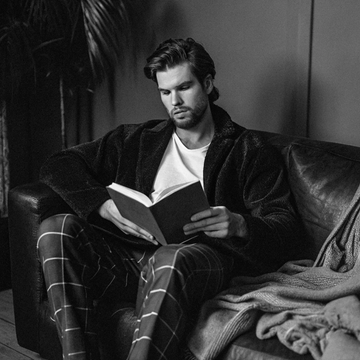 Homme lisant sur un canapé, vêtu d'un pyjama SLAZZ à carreaux et d'un peignoir noir, ambiance cosy en noir et blanc